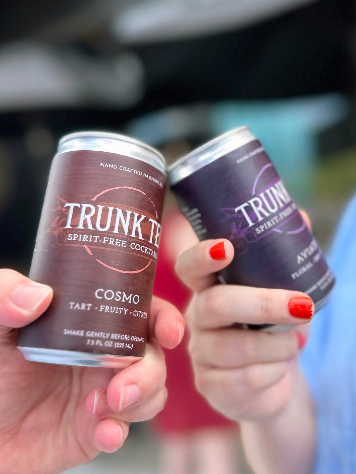 Two people touching two cans of Trunk Tea Spirit-Free Cocktails together, the cans are a Cosmo and a Aviation