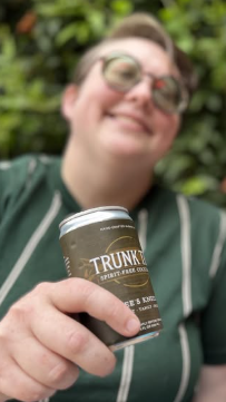 Emily Wines, founder of Trunk Tea Spirit-Free Cocktails  wearing a green shirt with stripes smiling and holding a can of Bee's Knees 