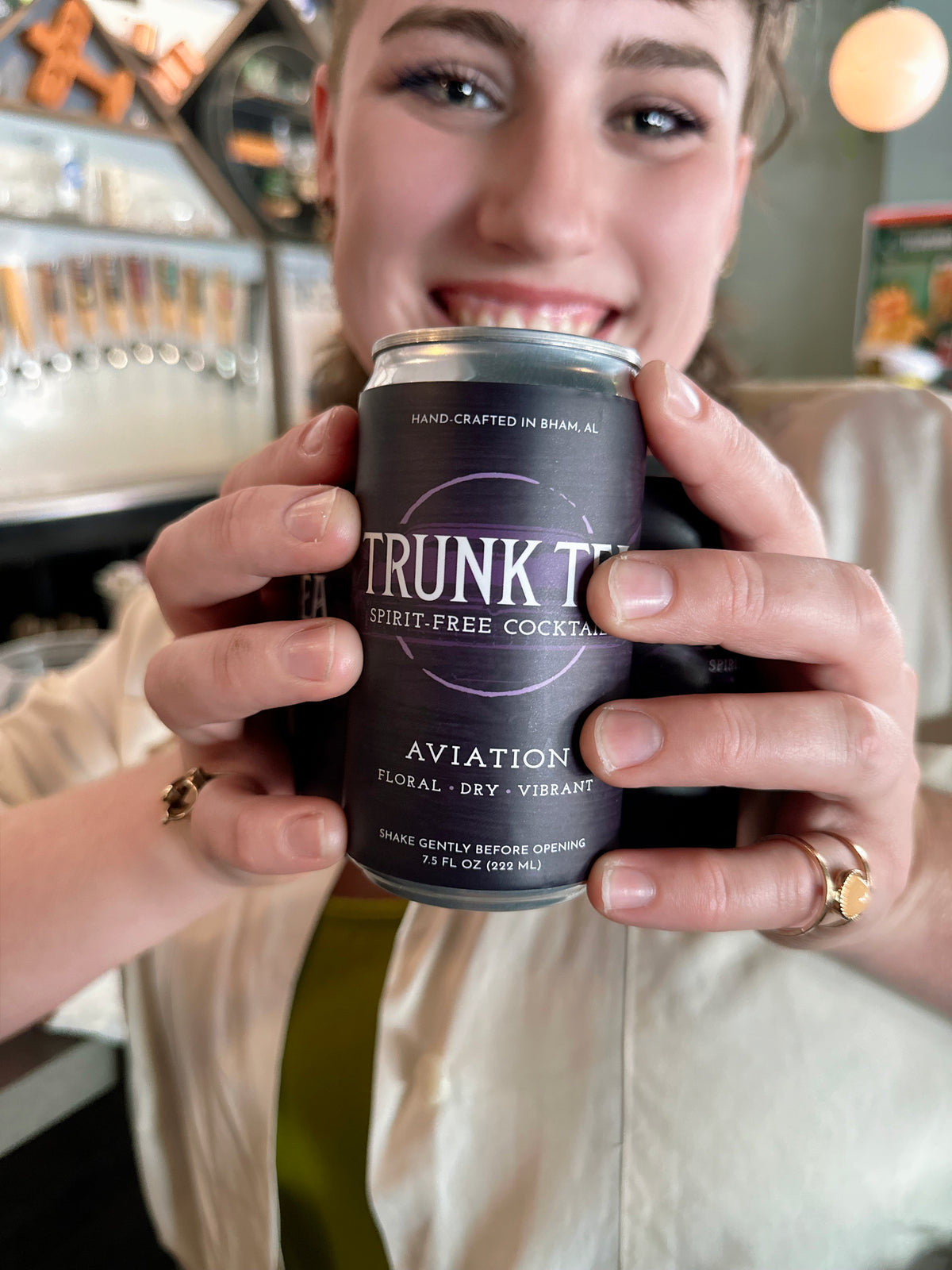 Smiling non-binary individual holding a can of Trunk Tea Spirit-Free Aviation  Mocktail between their hands with a bar in the background

