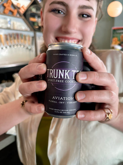 Smiling non-binary individual holding a can of Trunk Tea Spirit-Free Aviation  Mocktail between their hands with a bar in the background

