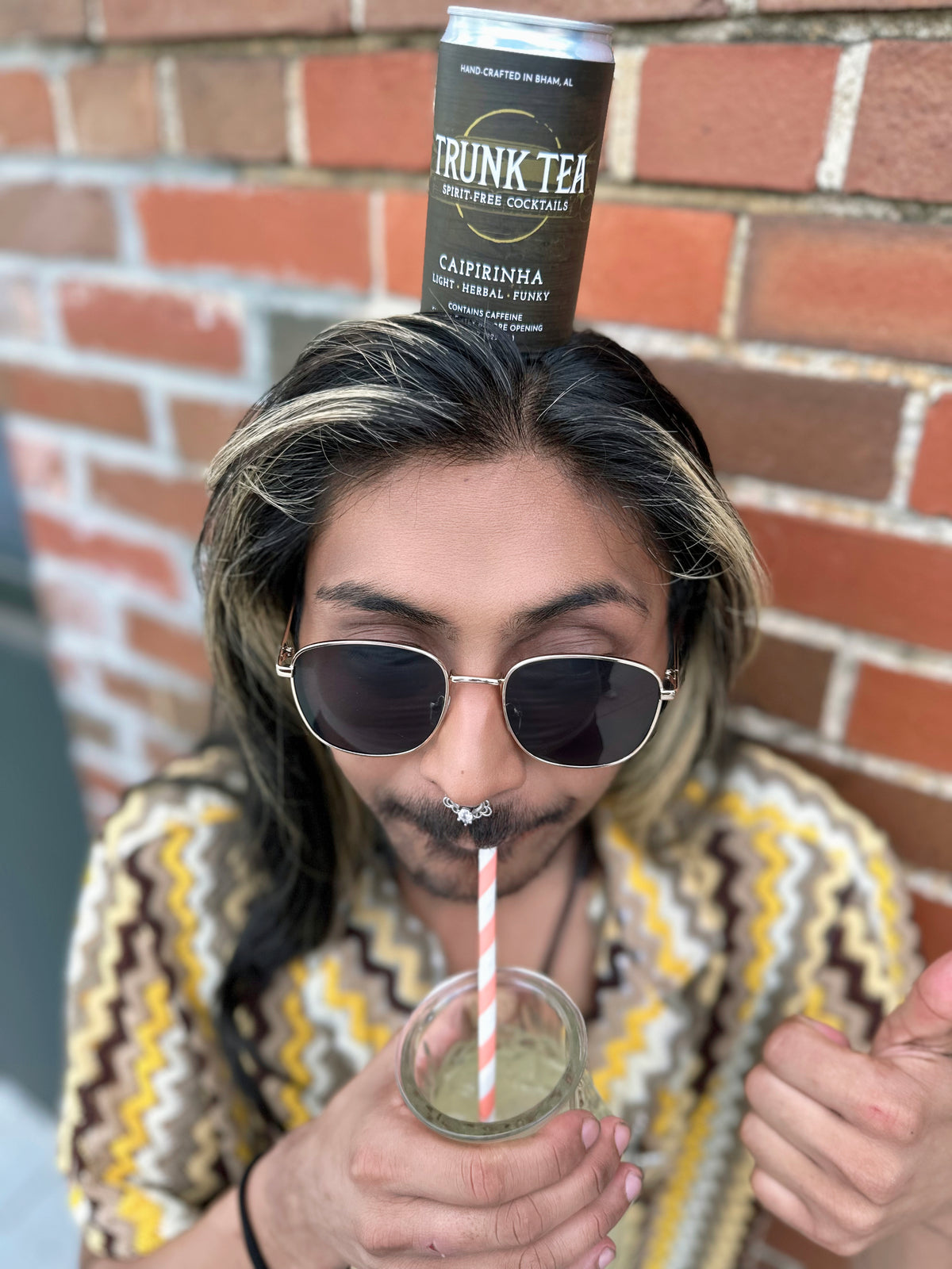 A can of Trunk Tea Spirit-Free Caipirinha Mocktail, balancing on non-binary individuals head, while drinking Caipirinha from a fun pineapple shaped glass. 
