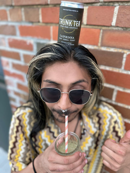 A can of Trunk Tea Spirit-Free Caipirinha Mocktail, balancing on non-binary individuals head, while drinking Caipirinha from a fun pineapple shaped glass. 
