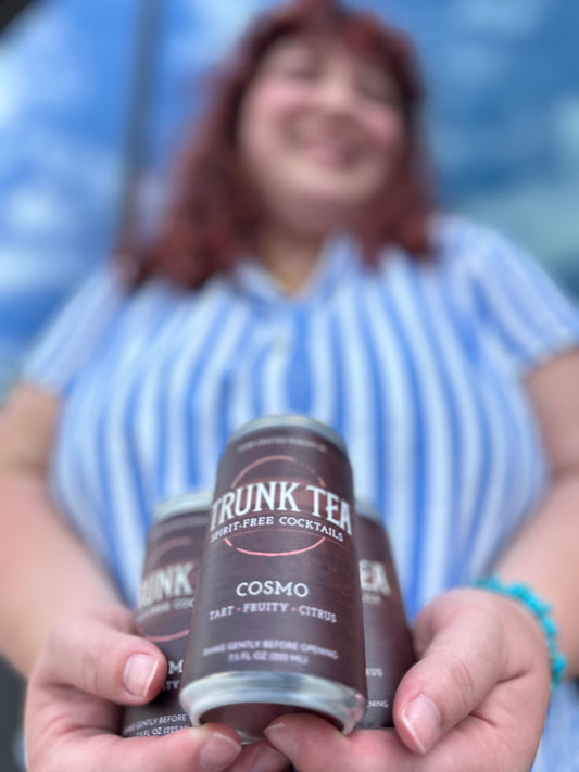 Red haired female in a blue and white striped blouse cradling three cans of Trunk Tea Spirit-Free Cosmo Mocktails in her hands
