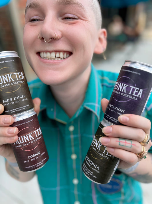 Image of smiling non-binary individual with shaved head holding the four flavors included in our Trunk Tea Spirit-Free Variety Pack. The Flavors are Aviation, Bee's Knees, Caipirinha and Cosmo 
